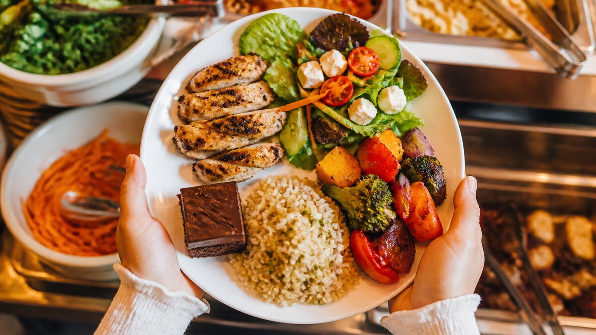 Two buffet plates with grilled meat, rice, roasted vegetables, salad, sauces, and a small dessert viewed from above.