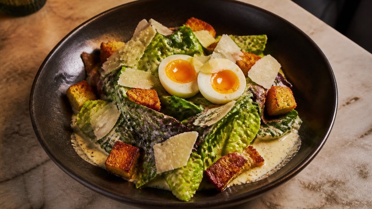 A restaurant salad with creamy dressing, parmesan, and croutons in a dark ceramic bowl on a marble table.